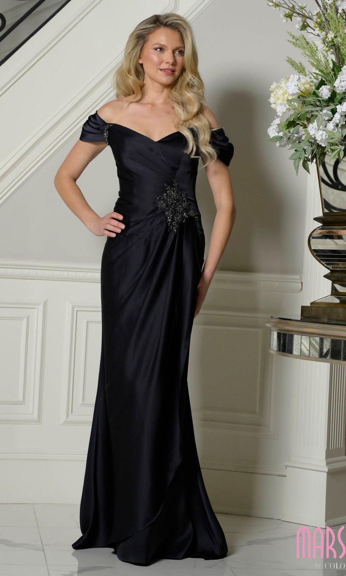A woman stands indoors in the Marsoni MV1351 Off-Shoulder Long Formal Dress in black, featuring a decorative waist brooch, next to a small table with a floral arrangement.